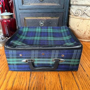 May include: A vintage plaid suitcase in shades of blue and green, with a black handle and trim. The suitcase has silver-toned hardware and a zipper closure. The suitcase is sitting on a wooden surface.