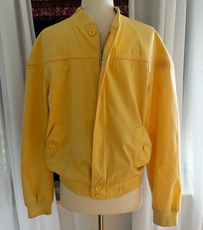 May include: A yellow bomber jacket with a zipper closure and button accents. The jacket has a ribbed collar and cuffs.