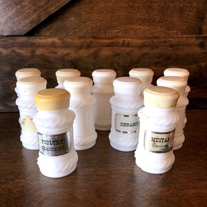 May include: Set of ten white milk glass spice jars with cork lids. The jars have a rope design and gold labels with the names of spices, including "Cinnamon", "Poultry Seasoning", and "Mustard Ground".