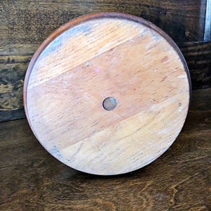 May include: The bottom of a wooden round cake stand with a hole in the center. The stand is made of light brown wood with a darker brown rim.