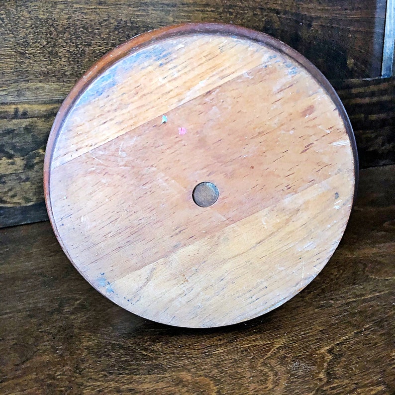 May include: The bottom of a wooden round cake stand with a hole in the center. The stand is made of light brown wood with a darker brown rim.