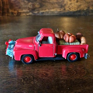Könnte beinhalten: Ein roter Spielzeug-Pickup mit einer Ladefläche voller Mini-Kürbisse und Heu. Der LKW hat einen blauen und weißen Weihnachtsbaumschmuck am vorderen Stoßfänger.