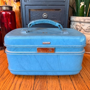 May include: A vintage blue suitcase with a handle and a metal clasp. The suitcase has a textured surface and a white trim. The handle is also blue. The suitcase is sitting on a wooden surface.