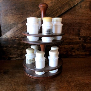 May include: A wooden spice rack with two tiers holds eight white milk glass spice jars with labels. The top tier has three jars, the bottom tier has five jars. The rack has a dark wood finish and a round base.