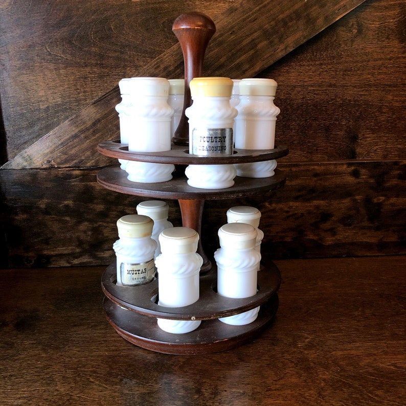 May include: A wooden spice rack with two tiers holds eight white milk glass spice jars with labels. The top tier has three jars, the bottom tier has five jars. The rack has a dark wood finish and a round base.