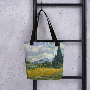 Op de afbeelding: Tote bag met een landschapsschilderij met een geel veld, groene bomen en een blauwe lucht met wolken. De tas heeft zwarte handvatten en hangt aan een zwarte metalen ladder.
