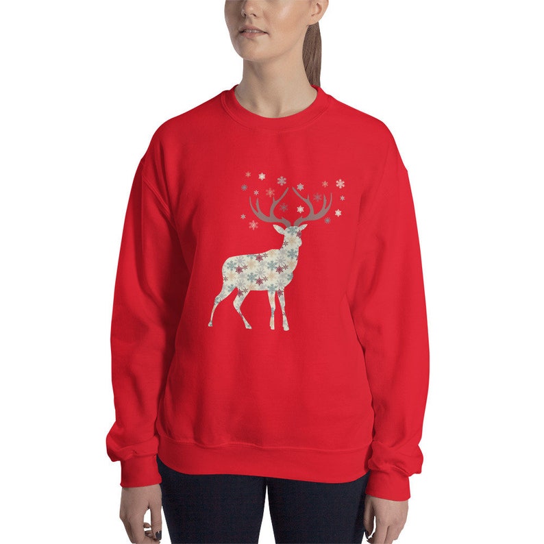 May include: Red sweatshirt with a white silhouette of a deer with snowflake patterned antlers and body. The deer is surrounded by small snowflakes.