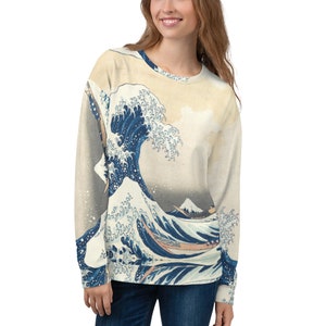 Pode incluir: Uma camisa de moletom branca com uma estampa azul e branca de uma grande onda e uma montanha no fundo. A estampa é uma reprodução de uma famosa gravura japonesa em madeira.