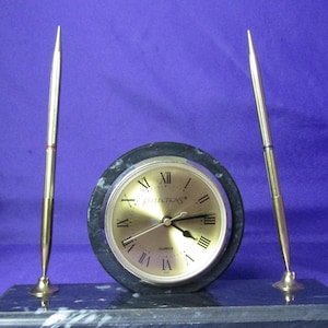 May include: A black marble desk clock with gold-tone hands and Roman numeral markings. The clock is on a black marble base with two gold-tone pen holders.
