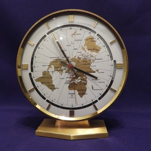 May include: A gold-toned world clock with a white face and black markings. The clock features a map of the world with major cities and time zones labeled. The clock is on a gold-toned stand.