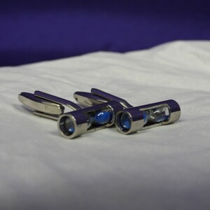May include: A set of three silver cufflinks with a small hourglass design. Each hourglass contains blue sand.