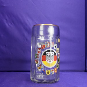 May include: A clear glass beer stein with a gold rim and a circular design featuring the coats of arms of the German states. The center of the design features the German flag with the words "Bundesrepublik Deutschland Germany".
