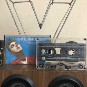May include: A cassette tape with a clear plastic case and a black label. The label has the text "MORRISSEY/BONA DRAG" and "PRODUCED BY STEPHEN STREET" and "PRODUCED BY CLIVE LANGER AND ALAN WINSTANLEY". The cassette tape is sitting on a wooden surface.