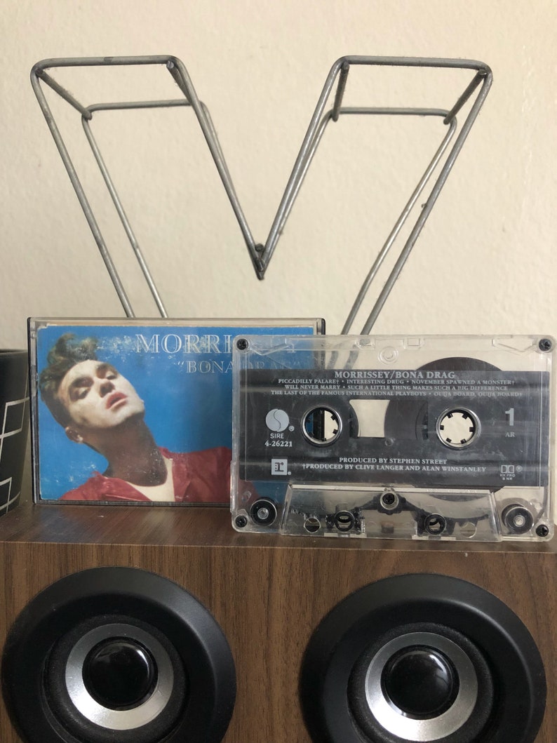 May include: A cassette tape with a clear plastic case and a black label. The label has the text "MORRISSEY/BONA DRAG" and "PRODUCED BY STEPHEN STREET" and "PRODUCED BY CLIVE LANGER AND ALAN WINSTANLEY". The cassette tape is sitting on a wooden surface.