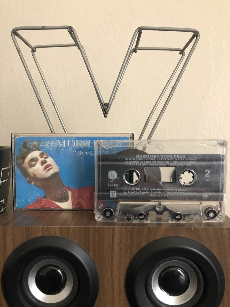 May include: A clear cassette tape with a black label. The label has the text "MORRISSEY/BONA DRAG" and the song titles "HAIRDRESSER ON FIRE", "EVERYDAY IS LIKE SUNDAY", "HE KNOWS I'D LOVE TO SEE HIM", "YESAAM BUNDE", "LUCKY LISP", "SEDEHEAD", and "LISAPPOINTE". The cassette tape is sitting on a wooden surface.