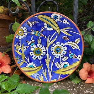 May include: A blue ceramic plate with a floral design. The plate has a white background with yellow, green, and blue flowers. The plate has a brown rim.