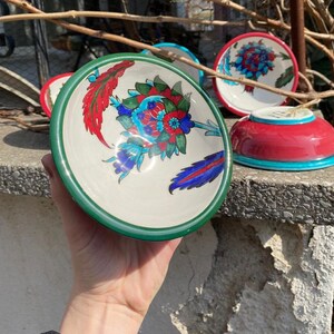 May include: A set of four hand-painted ceramic bowls with intricate floral designs in shades of blue, red, and green. The bowls have a white background and a green rim.