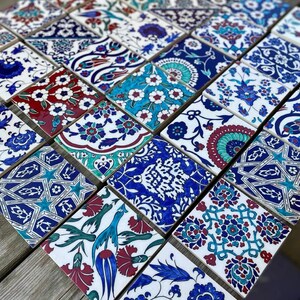 May include: A collection of 25 colorful ceramic tiles with intricate floral and geometric patterns in shades of blue, red, green, and white. The tiles are arranged in a grid pattern on a wooden surface.