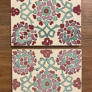 May include: Two square ceramic tiles with a white background and a repeating floral pattern in shades of red, purple, and green. The pattern features stylized flowers and leaves.