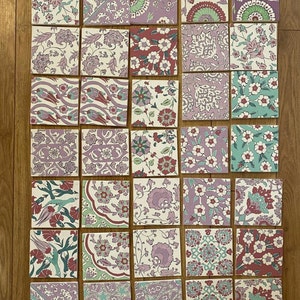 May include: A collection of 25 square ceramic tiles with intricate floral patterns in shades of pink, green, and blue. The tiles are arranged in a grid pattern on a wooden surface.