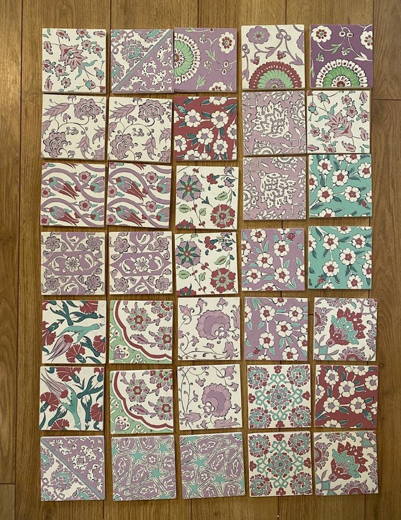 May include: A collection of 25 square ceramic tiles with intricate floral patterns in shades of pink, green, and blue. The tiles are arranged in a grid pattern on a wooden surface.