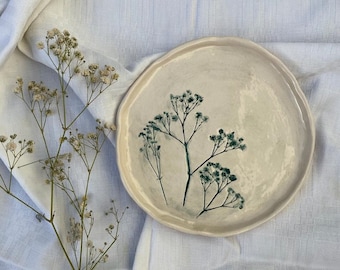 May include: A white ceramic plate with a delicate floral design in shades of green. The plate has a slightly irregular shape and a smooth, matte finish.