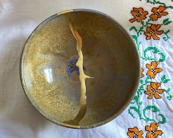 Puede incluir: Un cuenco de cerámica con un diseño de glaseado dorado. El cuenco tiene un glaseado moteado azul y marrón con una franja dorada en el centro.