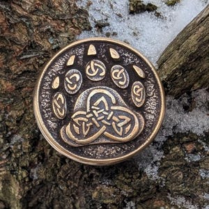 Könnte beinhalten: Ein runder, bronzefarbener Metallanhänger mit einem Bärenpfotenabdruck über einem keltischen Knotenmuster. Der Anhänger befindet sich vor dem Hintergrund eines verschneiten Astes.
