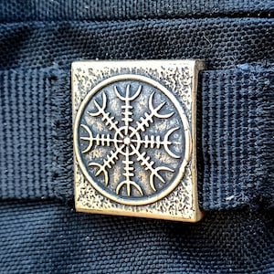 以下が含まれることがあります： 黒い布製のバッグのアップで、真鍮色の金属製のバックルが付いています。バックルには、8本のスポークと中央の円を持つ円形のデザインが施されています。デザインはコンパスや太陽を連想させます。