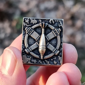 May include: A bronze-toned square metal pin with a military-themed design. The pin showcases a central projectile shape encircled by crossed elements within a circular frame. It has a textured, pixelated border, and is held in a hand.