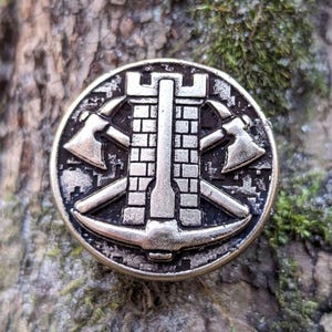 May include: A circular, antique silver-toned metal pin with a central tower design, two axes, and a pickaxe. The pin is set against a textured, mossy tree trunk background, creating a rustic aesthetic.