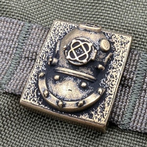 Può includere: Un ciondolo in ottone a forma di casco da sub con un design dettagliato. Il ciondolo è di forma quadrata e ha una superficie testurizzata.