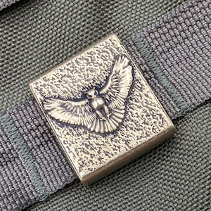 May include: A brass-colored metal buckle with a detailed design of a bird in flight. The buckle is attached to a green fabric strap.