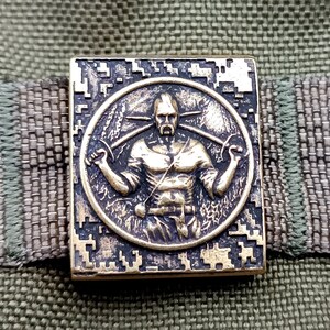 Op de afbeelding: Een bronzenkleurige metalen patch met een gedetailleerd ontwerp van een man die twee zwaarden vasthoudt. De man draagt ​​een tuniek en heeft een baard. De patch is omgeven door een vierkant frame met een gepixelde patroon.