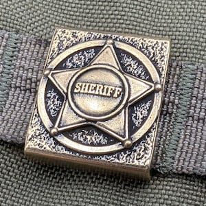 以下が含まれることがあります： 中央に「SHERIFF」の文字が書かれた、5つのポイントを持つ星形の金色の金属製の保安官バッジです。