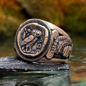 May include: A bronze-toned metal ring featuring a circular face with an embossed owl and the Greek letters Alpha, Omicron, and Sigma. The sides of the ring display a detailed relief of a Spartan helmet with a crest, set against a dark background.