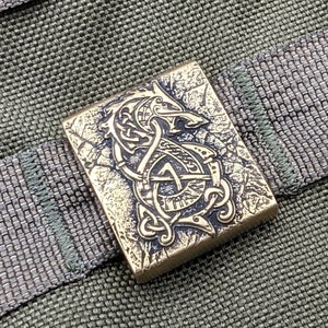 Peut inclure: Un carré en métal doré avec un motif détaillé de dragon et de nœuds. Le carré est sur un fond de tissu vert.