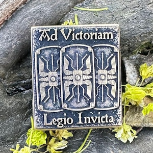 May include: A silver-coloured metal plaque with the words "Ad Victoriam" and "Legio Invicta" in decorative lettering. The plaque features three shield designs. The background is a natural wood surface with small yellow flowers.