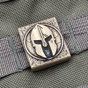 May include: A bronze-colored metal square patch with a circular design featuring a Spartan helmet in the center. The helmet is detailed with a raised design and a textured surface.