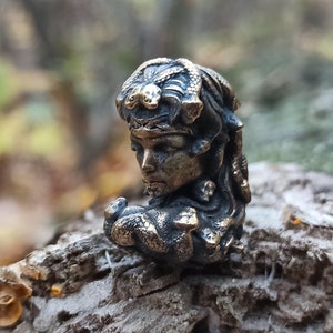 Puede incluir: Una figurita de bronce de una mujer con serpientes en el pelo. La figurita es detallada y tiene un aspecto realista. La figurita est&aacute; sentada sobre un trozo de madera.