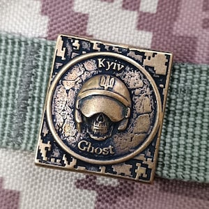 May include: A bronze-colored metal pin with a skull and helmet design. The pin has the text "Ghost" and "Kyiv" on it, along with the Ukrainian trident symbol.