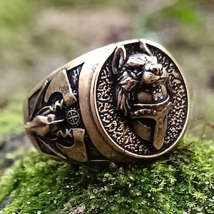 May include: A bronze ring with an intricate design featuring a wolf's head and a cross. The ring is set against a background of green moss.