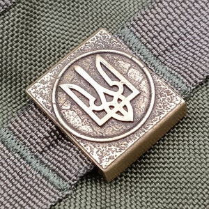 May include: A close-up of a bronze-colored metal patch with a stylized trident design. The patch is attached to a green and gray fabric strap.