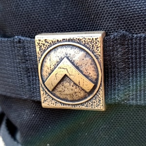 以下が含まれることがあります： 黒いバックパックのアップで、ゴールドの金属製のバックルに、シェブロンのデザインが施されたスタイリッシュなシールドが描かれています。