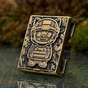 May include: A rectangular brass-toned metal bead featuring a raised relief design of a cat wearing a tactical helmet and vest. The background of the bead has a textured, pixelated pattern, and the sides include a vertical slot for cord threading.