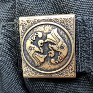 May include: A close-up of a gold-coloured metal badge featuring a detailed design of two stylised birds facing each other. The badge is attached to a black fabric surface.