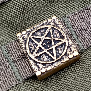 Peut inclure: Un charme pentagramme de couleur laiton avec une surface texturée. Le charme est attaché à une sangle en tissu brun et vert.
