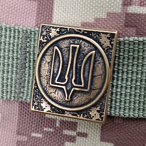 May include: A bronze-colored metal pin with a circular design featuring a stylized trident, the national symbol of Ukraine.