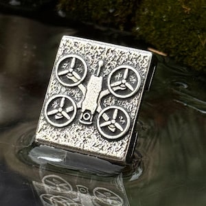 Peut inclure: Pendentif argenté rectangulaire avec un motif de drone. Il présente quatre hélices et un corps central. Le pendentif se reflète dans l'eau. Le fond montre de la mousse verte.