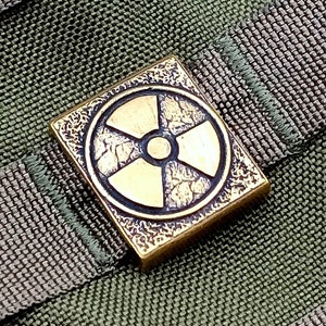 May include: A brass-colored square-shaped buckle with a raised, textured, circular design of a radiation symbol. The buckle is attached to a green and gray fabric background.